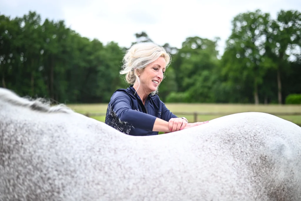 Osteopaat Sabrina Barmentloo voert een osteopathische behandeling uit op een wit paard in een groene buitenomgeving. Ze lacht terwijl ze haar handen op de rug van het paard legt, wat een rustige en ontspannen sfeer weerspiegelt