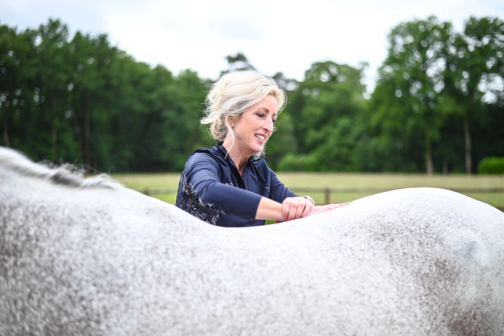Osteopaat Sabrina Barmentloo voert een osteopathische behandeling uit op een wit paard in een groene buitenomgeving. Ze lacht terwijl ze haar handen op de rug van het paard legt, wat een rustige en ontspannen sfeer weerspiegelt