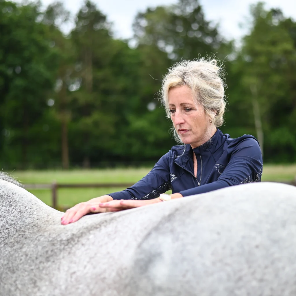 Sabrina Barmentloo van 'Osteopaat voor je Paard' voert een zorgvuldige inspectie uit op de rug van een wit paard. Deze afbeelding illustreert een blog over de anatomische overeenkomsten en verschillen tussen mensen en paarden, met nadruk op skeletstructuur, spijsverteringsstelsel, zintuigen, ademhalingsstelsel, en functionele aanpassingen zoals beweging, snelheid, voeding, en communicatie.