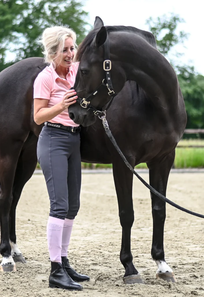 Sabrina Barmentloo van 'Osteopaat voor je Paard' staat lachend naast een zwart paard tijdens een cranio-sacraal behandeling. Deze afbeelding illustreert de zachte en gerichte aanpak van het behandelen van het cranio-sacraal systeem bij paarden om hun algehele welzijn te verbeteren.