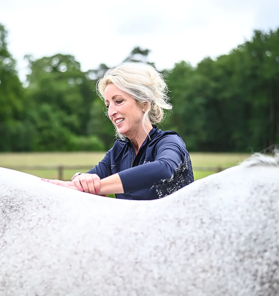 Sabrina Barmentloo van 'Osteopaat voor je Paard' voert een osteopathische behandeling uit op de rug van een wit paard in een rustige buitenomgeving. Deze afbeelding illustreert haar brede expertise in paardenbehandeling, waaronder sportmassage, cranio sacraal technieken, de Masterson methode, neuro myofasciale release, medical taping, dry needling, en kruidengeneeskunde.