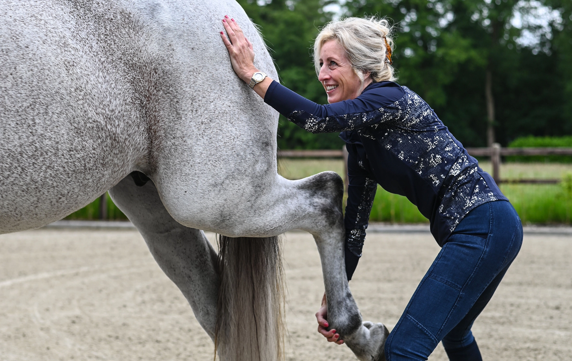 Sabrina Barmentloo, osteopaat bij 'Osteopaat voor je Paard', voert een mobiliteitsoefening uit op het achterbeen van een wit paard. Haar brede glimlach toont haar toewijding en plezier in het werken met paarden. Sabrina is een ervaren osteopaat met opleidingen in onder andere paardenosteopathie, sportmassage en cranio sacraal technieken.