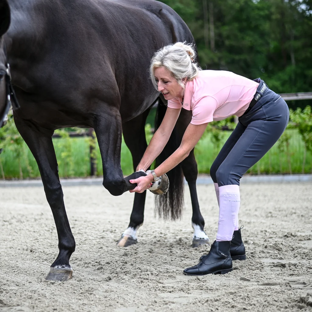 Sabrina Barmentloo van 'Osteopaat voor je Paard' voert een mobiliteitsoefening uit op het been van een zwart paard. De afbeelding biedt inzicht in standsafwijkingen en bewegingsrichtingen bij paarden, met uitleg over veelvoorkomende afwijkingen zoals nauw, bodemnauw, wijd, en de biomechanische gevolgen hiervan.